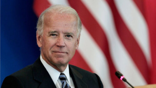 President Biden in a suit and tie stands at a podium, holding a microphone and delivering a speech. He has short hair and is focused on the audience, with the flag of the United States visible in the background. The setting is formal, indicating an official event or press conference.