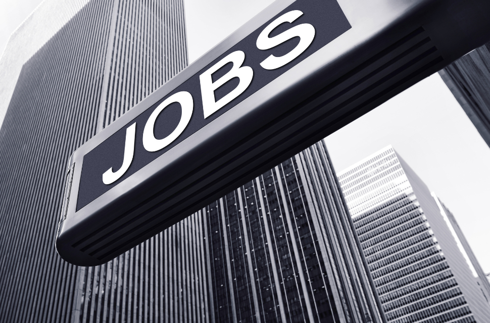 A black and white photo of a "JOBS" sign hanging in front of several tall office buildings, symbolizing June employment in an urban corporate environment. The buildings have a modern architectural design and the sky is clear.