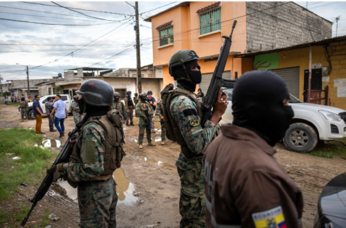 Ecadorian armed security forces in camouflaged uniform standing guard on a street with onlookers in the background.