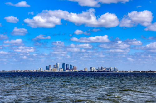 A panoramic view of a city skyline under a vast blue sky dotted with cumulus clouds, as seen from across the rippling waters of a large body of water, now threatened by rising tides.