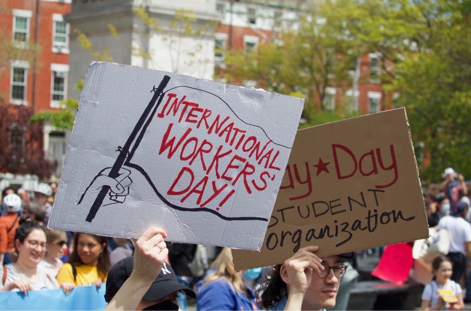 Protestors holding signs, one reading "International Workers Day!" with a graphic of a raised fist symbolizing unions, and the other saying "May Day student organization," at a rally. Photo: Pamela Drew, NYC May Day Workers March.