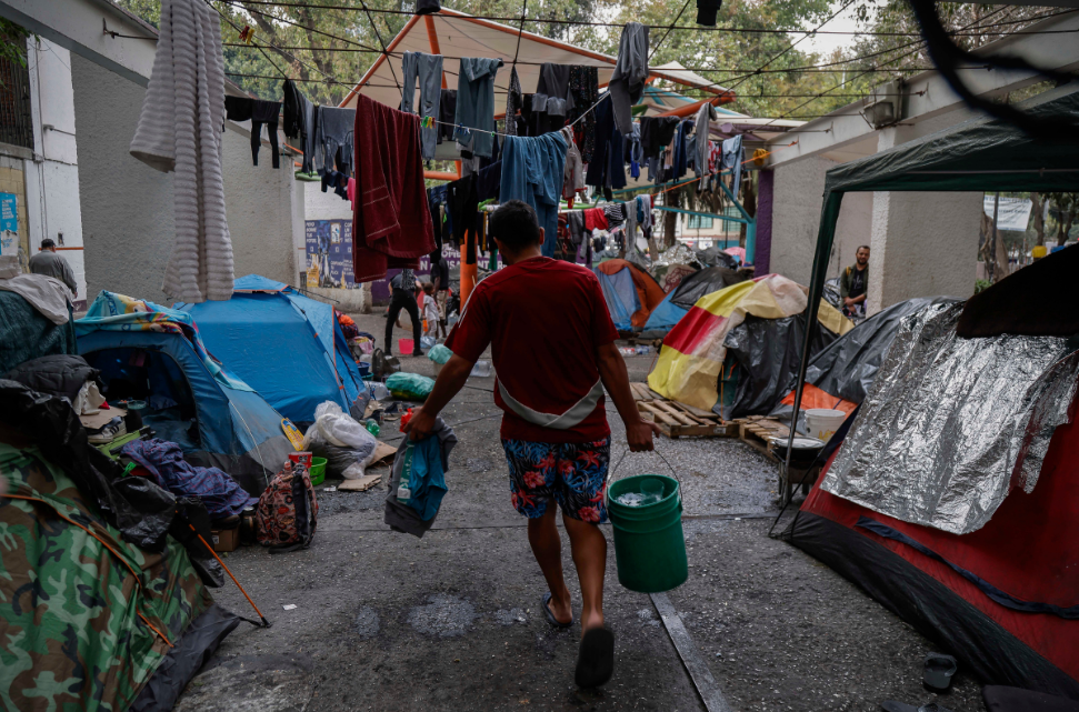 A man carries a bucket through a makeshift camp with tents and hanging laundry in an urban area, depicting a scene of temporary housing or a refugee settlement impacted by poverty due to sanctions.
