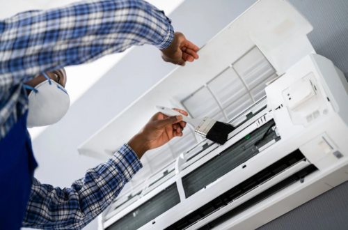 A technician in a mask and blue coveralls uses a brush to improve the indoor air quality by cleaning the inside components of a white air conditioning unit.