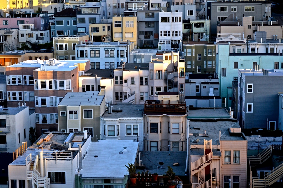 A densely packed urban neighborhood with various multi-story rental buildings in pastel colors including beige, light blue, and yellow. Structures feature flat and gabled roofs, balconies, and windows. The rooftops have visible antennas, solar panels, and vents—a typical scene for renters struggling to find affordable housing.