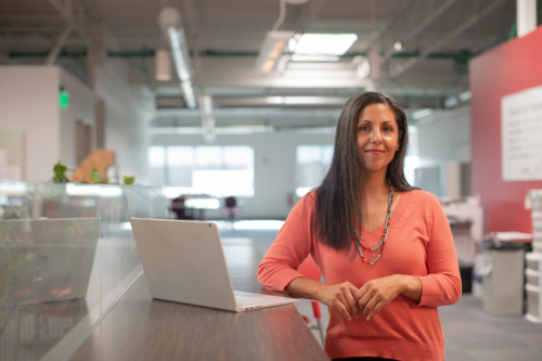 A confident working mother in a coral top stands at a counter with a laptop in a modern office, looking at the camera. Historically women’s poverty rates have been higher than men’s rates.