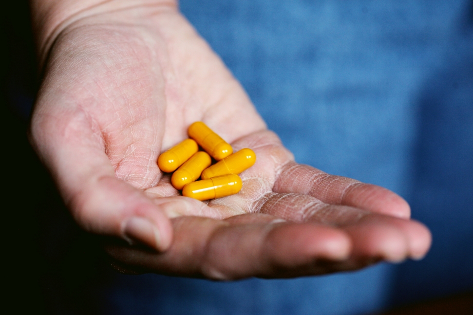 A close-up of an open hand holding several yellow capsules, emphasizing concerns about the impact of private equity in healthcare. The person appears to be a doctor wearing a blue garment, with the focus on the palm and capsules, set against a blurred background.