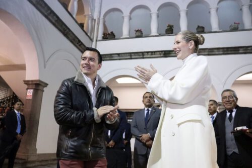 A dark-haired young man in a dark leather jacket and a woman with blond hair in a white coat stand in the foreground. Several men in suits stand in the background, in a plaza.