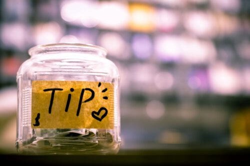 A glass tip jar with paper currency and coins inside is placed on a counter, indicating gratuities. The jar has a hand-drawn label with the word 