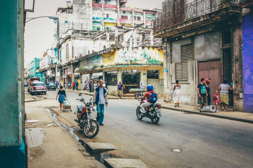 A vibrant street scene in a dilapidated urban area with weathered, run-down buildings. People walk and socialize while scooters and motorcycles pass by. The atmosphere is lively, yet the buildings are showing signs of age, decay and, neglect, the effect of economic sanctions.
