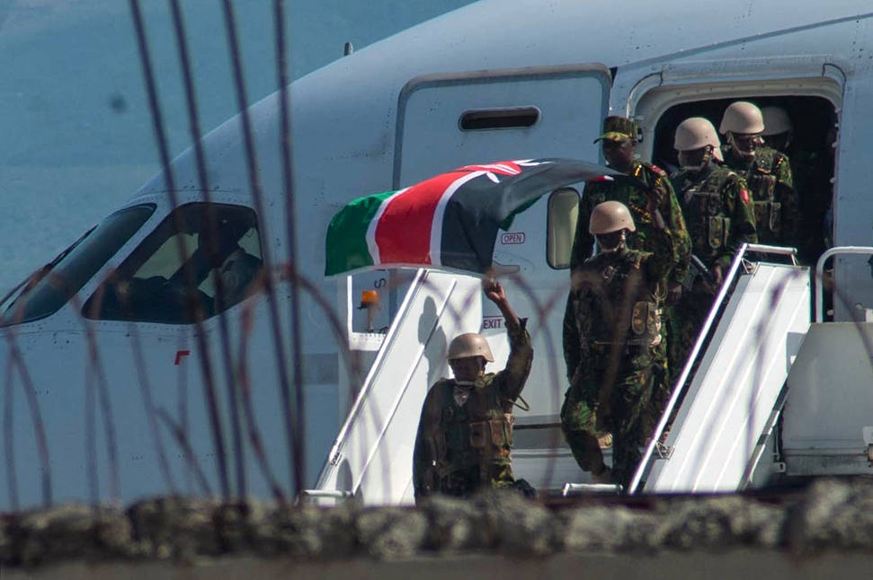 A group of uniformed soldiers are seen descending from an airplane. One soldier in front waves the Kenyan flag. The Kenyan forces, wearing helmets and face masks, are emerging from the aircraft onto a staircase upon their arrival in Haiti. Credit: CLARENS SIFFROY.