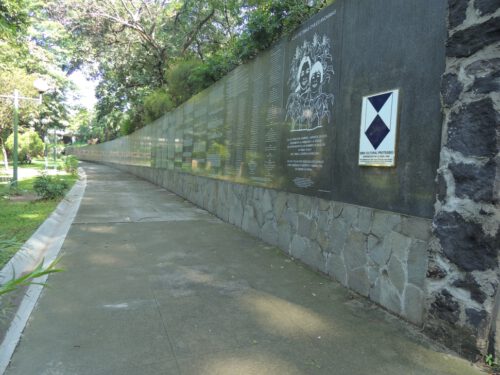 human-rights-torn-between-justice-for-el-salvadors-war-victims-and-u-s-complicity-A long memorial wall displaying various texts and images stretches along a shaded sidewalk. The path is surrounded by greenery and lawned areas. A blue and white informational plaque is mounted on a stone section of the wall near the foreground. Maxu rm, CC BY-SA 3.0 , via Wikimedia Commons