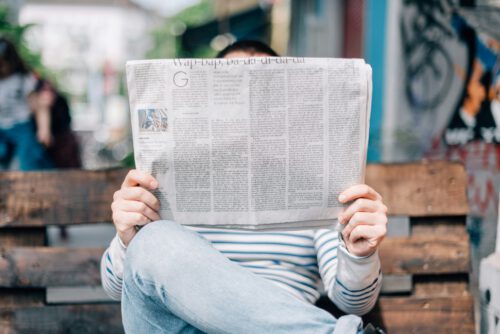 A person in a striped shirt is sitting on a wooden bench, holding and possibly reading a copy of the Washington Post. Their face is not visible as the newspaper covers it entirely. The background is slightly blurred, showing some wall art and a person in the distance.