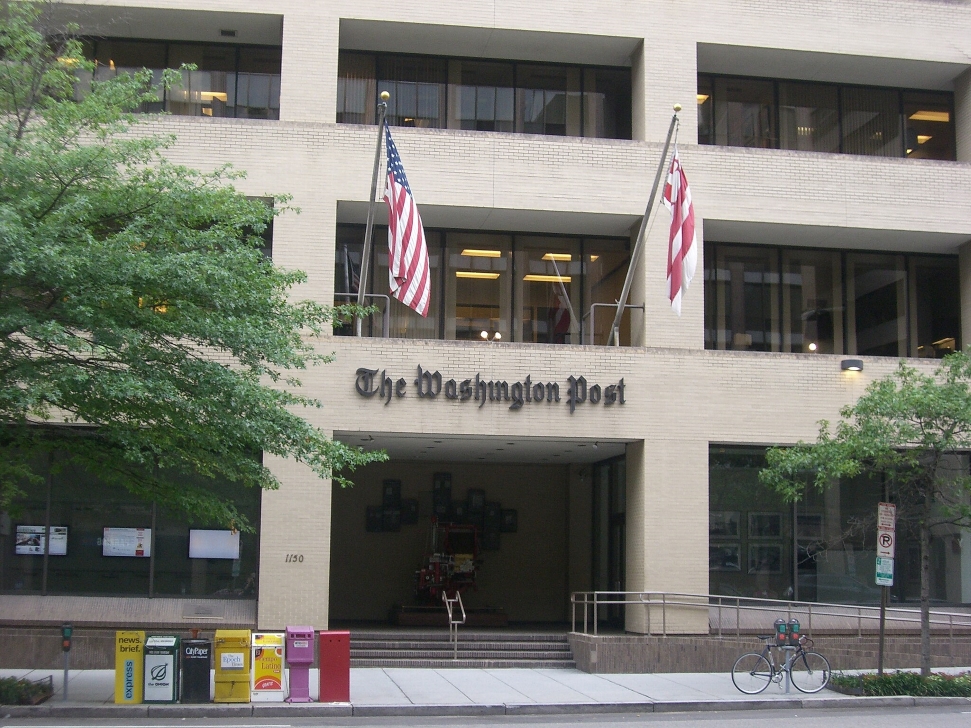 The front facade of The Washington Post building is shown with its name displayed above the entrance. Two flags, one American and one with red and white bars, are flying next to the entrance. Newspaper vending machines and a bicycle are visible on the sidewalk.