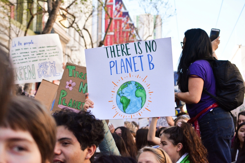 A crowd of people at a climate change and extreme weather protest in the south. The focal point is a sign held by a person in a purple shirt reading "THERE IS NO PLANET B," with an illustration of Earth. Other signs and protesters with various messages are in the background.