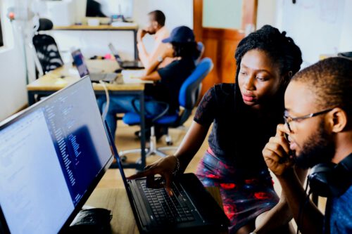 Two people are working together at a computer desk, looking at code on a monitor. The woman is pointing at the screen while the man observes. In the background, other individuals engaged in computer work contribute to fostering black employment in this diverse shared office space.