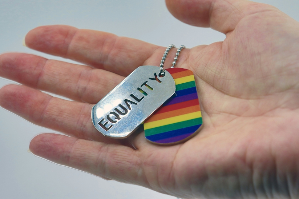 A hand is holding two dog tags: one is metallic with the word "EQUALITY" engraved on it, and the other is brightly colored with rainbow pride stripes. The image symbolizes support for LGBTQ+ workers and their rights to equality.