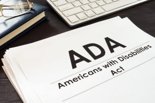 A close-up image of a document titled ADA Americans with Disabilities Act on an office desk. The desk also holds a part of a white computer keyboard and a blue notepad. The document's title is prominently displayed in bold text.