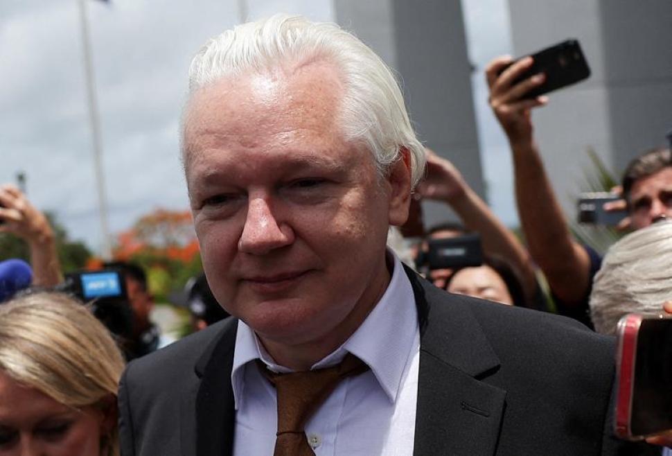 Julian Assange, wearing a suit and tie, is surrounded by people taking photos and videos with their phones. The scene could be outdoors at a significant location, possibly capturing a momentous event involving Julian Assange. Blurred figures and building structures form the background.