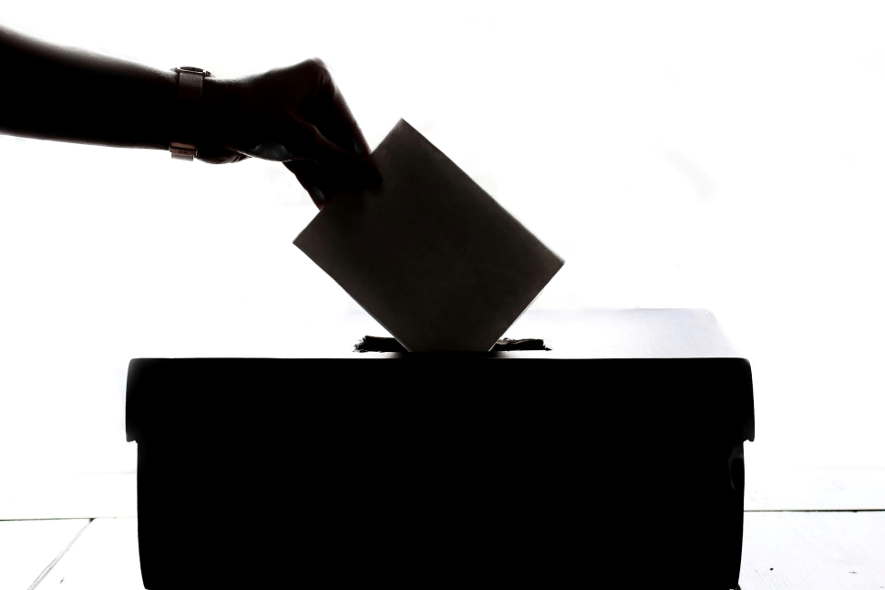 A silhouette of a hand placing a ballot into a ballot box, symbolizing the importance of the economy in the November elections. The background is white, with subtle details visible on the hand, including a wristwatch. The ballot box is dark and rectangular.
