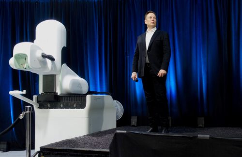 Elon Musk, in a black suit, stands on a stage next to a large white machine against a backdrop of dark blue curtains. He is holding a microphone in one hand and looking to the side.