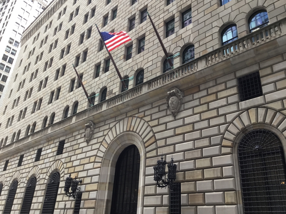 The Federal Reserve Bank in New York with an American flag flying above its entrance stands imposing. The facade features arched windows and decorative stonework. Numerous windows are visible on the upper floors, and three flagpoles are mounted between them, echoing discussions that the federal reserve must raise rates.