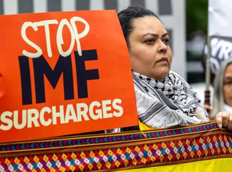 A person stands holding a bright orange sign that reads "STOP IMF SURCHARGES" in bold black text. They look determined and wear a patterned scarf. The edge of a multicolored woven fabric is visible in the foreground. Other blurred figures are in the background.