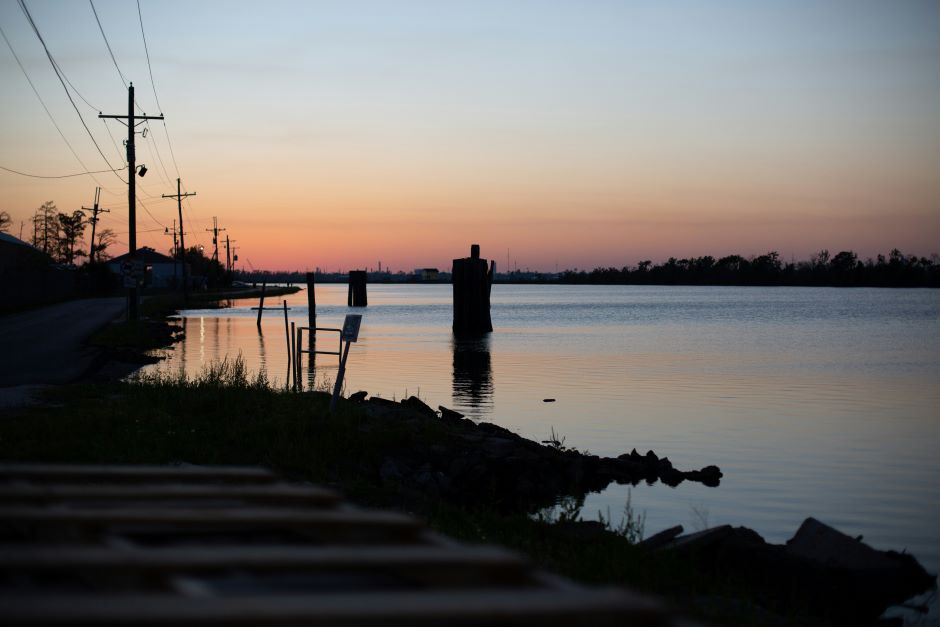 A tranquil scene at dusk with a calm water body reflecting the fading light of the setting sun. Silhouettes of trees, utility poles, and dock structures are visible along the shore, embodying efforts in saving coastal Louisiana. The sky displays a gradient of colors from orange and pink to deep blue.