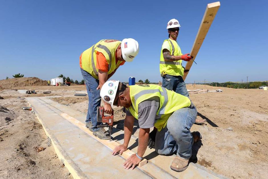 Three construction workers in safety vests and helmets work at a construction site. One person is drilling into a wooden plank while kneeling, another holds a plank in place, and the third stands holding a long piece of wood amidst sand and equipment. Will new OSHA Heat Standards change how outdoor work is done?