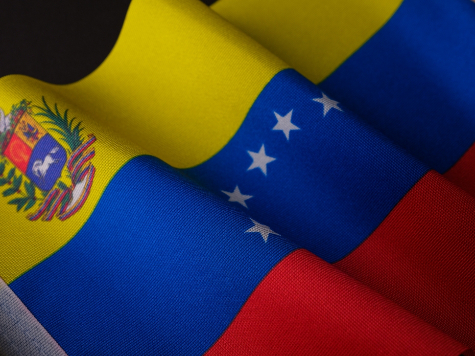 Close-up of the Venezuelan flag, displaying its yellow, blue, and red horizontal stripes. The blue stripe features eight white stars forming an arc, and the yellow stripe contains a national coat of arms in the upper left corner. Elections.