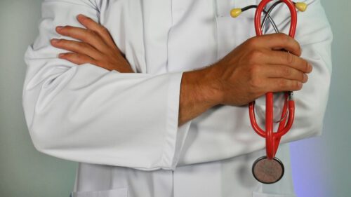 A person in a white lab coat stands with their arms crossed, holding a red stethoscope in one hand, representing the fight over private equity and health inequity.