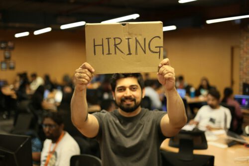 A man stands in an office filled with people working at desks, holding up a cardboard sign with the word 