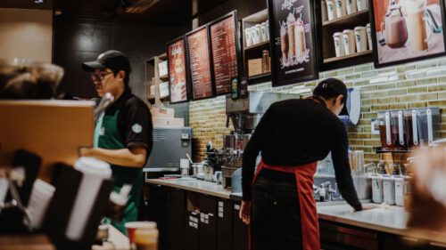 Two baristas earning minimum wage are working behind the counter in a coffee shop. One, wearing a black shirt and green apron, operates a coffee machine, while the other, in a red apron, cleans the counter. The menu boards above them display various drink options.