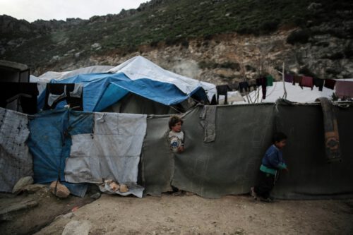 Two children stand near makeshift tents with tarps and cloths in front of a rocky hillside. The shelters appear worn, and clothes are hung on a line above. The landscape is rugged with vegetation scattered across the hills. The mood is somber, reflecting poverty as a result of sanctions.