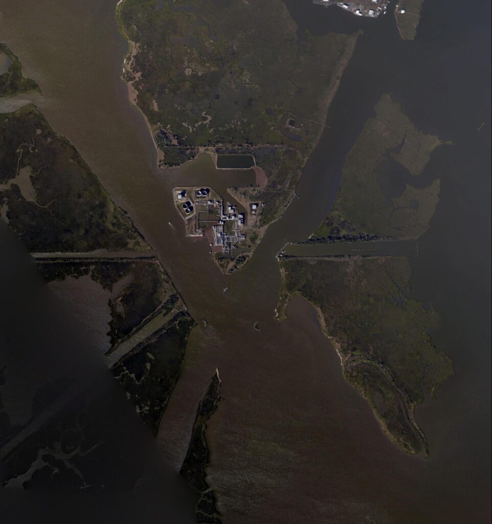 An aerial view of an industrial complex located on a narrow peninsula surrounded by water. The complex is connected by several roads and bridges to the surrounding land. The surrounding area appears largely undeveloped, with wetlands and natural landscapes. An aerial view of an industrial complex located on a narrow peninsula surrounded by water. Illustrating why efforts are being made in saving coastal Louisiana. 