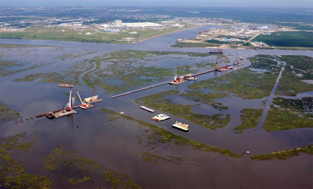 Aerial view of a marshland construction site showing cranes, barges, and a long bridge or pipeline being built over the water. The background features industrial buildings and infrastructure, all part of the effort toward saving coastal Louisiana adjacent to the expansive wetland area.