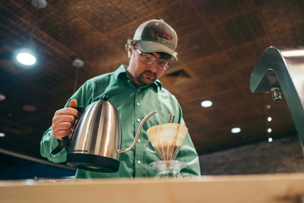 A low-wage worker wearing a green shirt and a cap is preparing coffee using a pour-over method. They are holding a gooseneck kettle, pouring hot water into a coffee dripper with ground coffee and a filter.