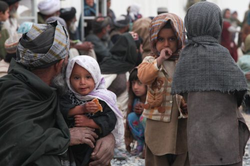 Two small Afghan children, dressed in traditional clothes, in the foreground look at the camera, as a third child in the background crouches, also looking at the camera. An older man holding one of the small children is turned away from the camera.