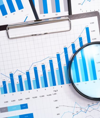 A collection of printed charts and graphs displaying blue and gray bar and line graphs on paper. A magnifying glass is placed on one sheet, highlighting the detailed statistical data bytes and figures on a clipboard.