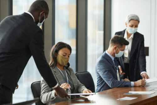 Four businesspeople wearing face masks are seated in a conference room, engaged in a meeting. One person is standing and handing a document to a seated colleague, while the two others review paperwork at the table. The backdrop of large windows hints at new norms in the era of Long Covid.