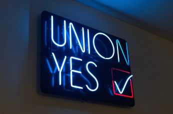 A neon sign on a wall reads "UNION YES" in blue letters, with a red checkmark inside a red square next to the word "YES". The sign glows brightly against a dark background.