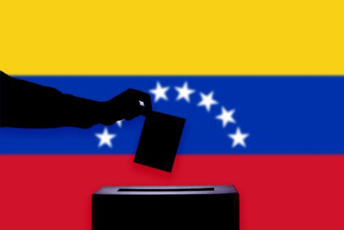 A silhouette of a hand casting a vote into a ballot box is depicted in front of the Venezuelan flag, which features yellow, blue, and red horizontal stripes with white stars forming an arc in the center. Venezuela election.