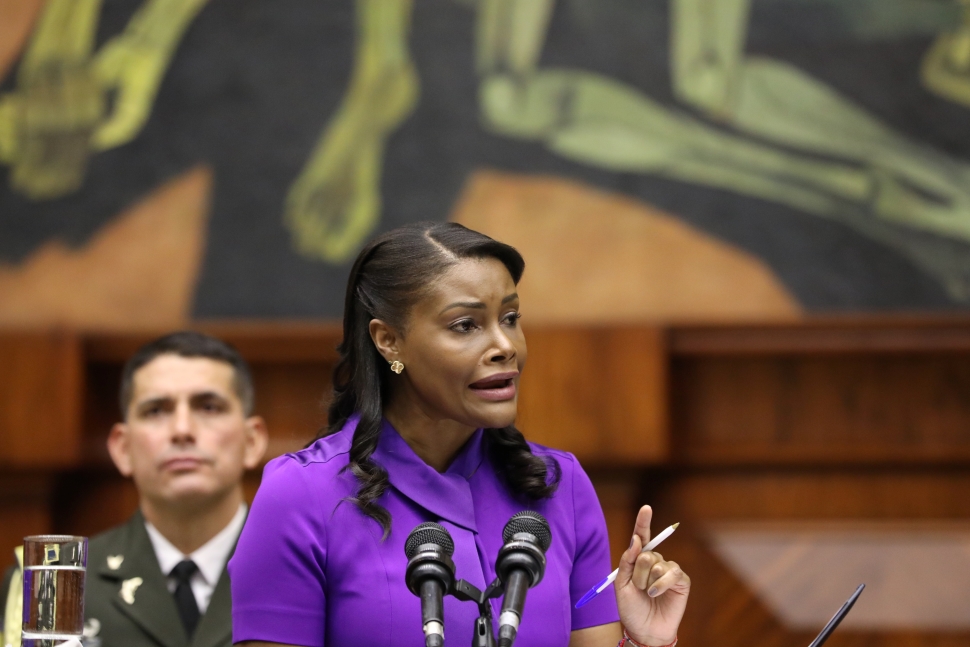 Ecuador News Round Up: Diana Salazar, in a purple dress, speaks at a podium with multiple microphones. Behind her, a man in a military uniform listens attentively. The background features an abstract mural with green and yellow hues.