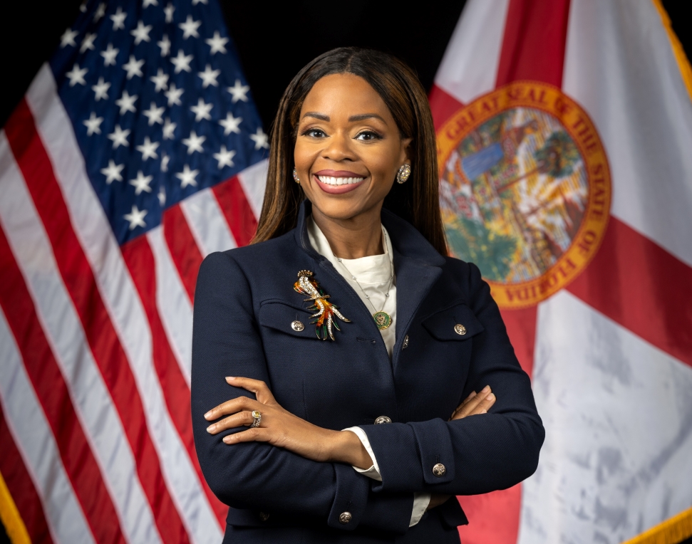 Rep. Sheila Cherfilus-McCormick 118th Congress. tands confidently with her arms crossed, wearing a dark blazer with buttons and a brooch. She smiles broadly, and behind her are the United States flag and the Florida state flag.