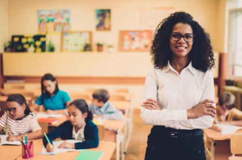 Despite the widening teacher pay gap, a smiling teacher with curly hair and glasses stands confidently in front of a classroom. Several young students, seated at desks, are engaged in writing and drawing activities. The classroom walls display colorful artwork and educational posters.