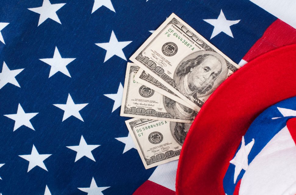 Four one-hundred-dollar bills partially tucked into a red hat, set against a backdrop of an American flag with white stars on a blue field. The composition suggests a theme of American patriotism and financial concepts, reflecting the idea that workers are better off in a thriving economy.