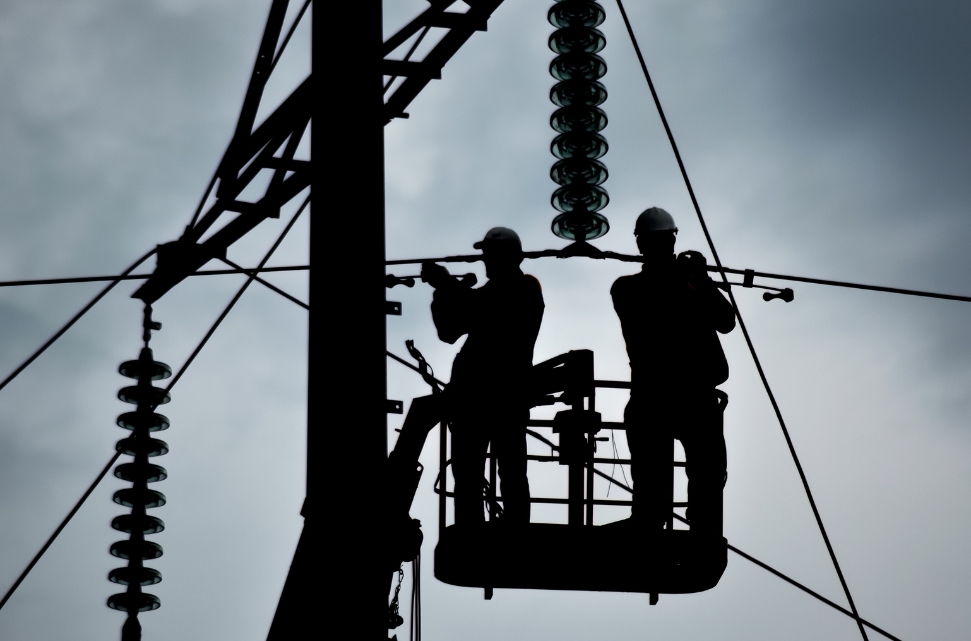 Silhouetted image of two electrcitity workers atop a raised platform, working on power lines against a cloudy West Virginia sky. Both are wearing hard hats and safety gear while installing or repairing components on a tall power pole.