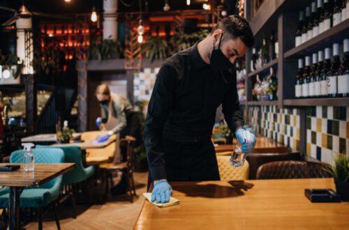 A man in a mask and gloves cleans a wooden table with a spray bottle and cloth in a restaurant, while a woman in the background also cleans. The restaurant has a modern design with teal chairs and a display of wine bottles.