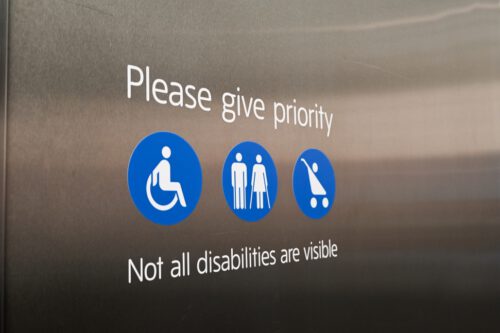 Sign on a metallic surface indicating priority for certain individuals. Icons represent a person in wheelchair, elderly people with canes, and a person with a stroller. Below, text reads: 