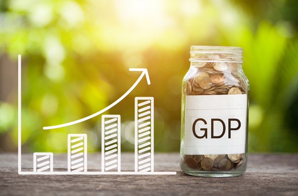 A glass jar labeled "GDP" filled with coins is placed on a wooden surface. Next to it, a graph with an upward trend arrow symbolizes economic growth. The background is blurred, showing greenery and sunlight.