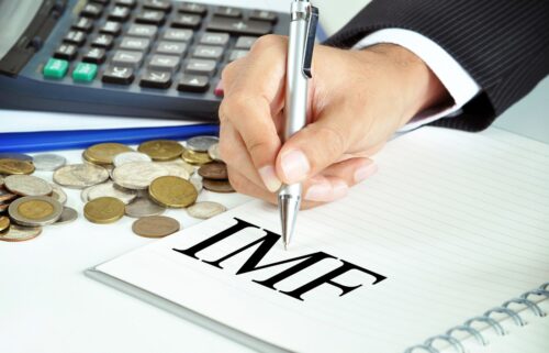 A person writing on a piece of paper IMF in big, bold, black letters with coins scattered around, in an indoor setting.